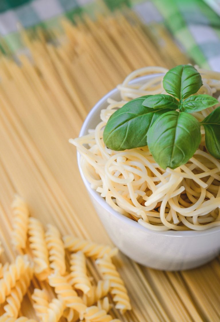 Creamy Pesto Pasta: A Harmony of Freshness and Indulgence 2 Bowl of cooked spaghetti with fresh basil, surrounded by raw spaghetti and fusilli pasta on a kitchen table.