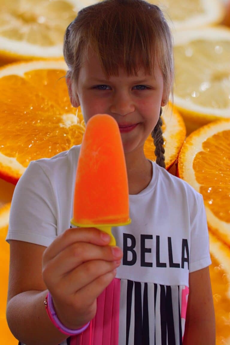 Child holding homemade orange popsicle with fresh orange slices background